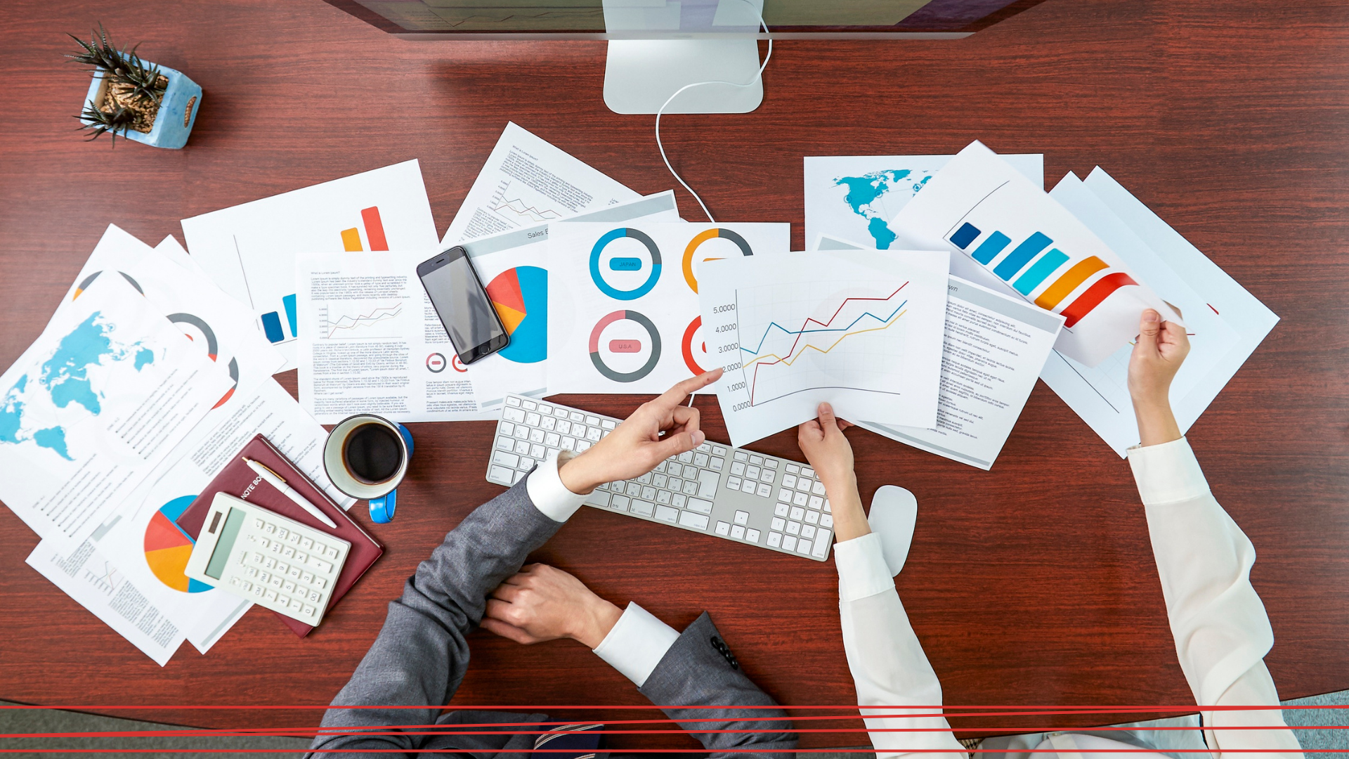 Due persone lavorano a una scrivania piena di grafici aziendali, diagrammi e una tastiera, viste dall'alto.