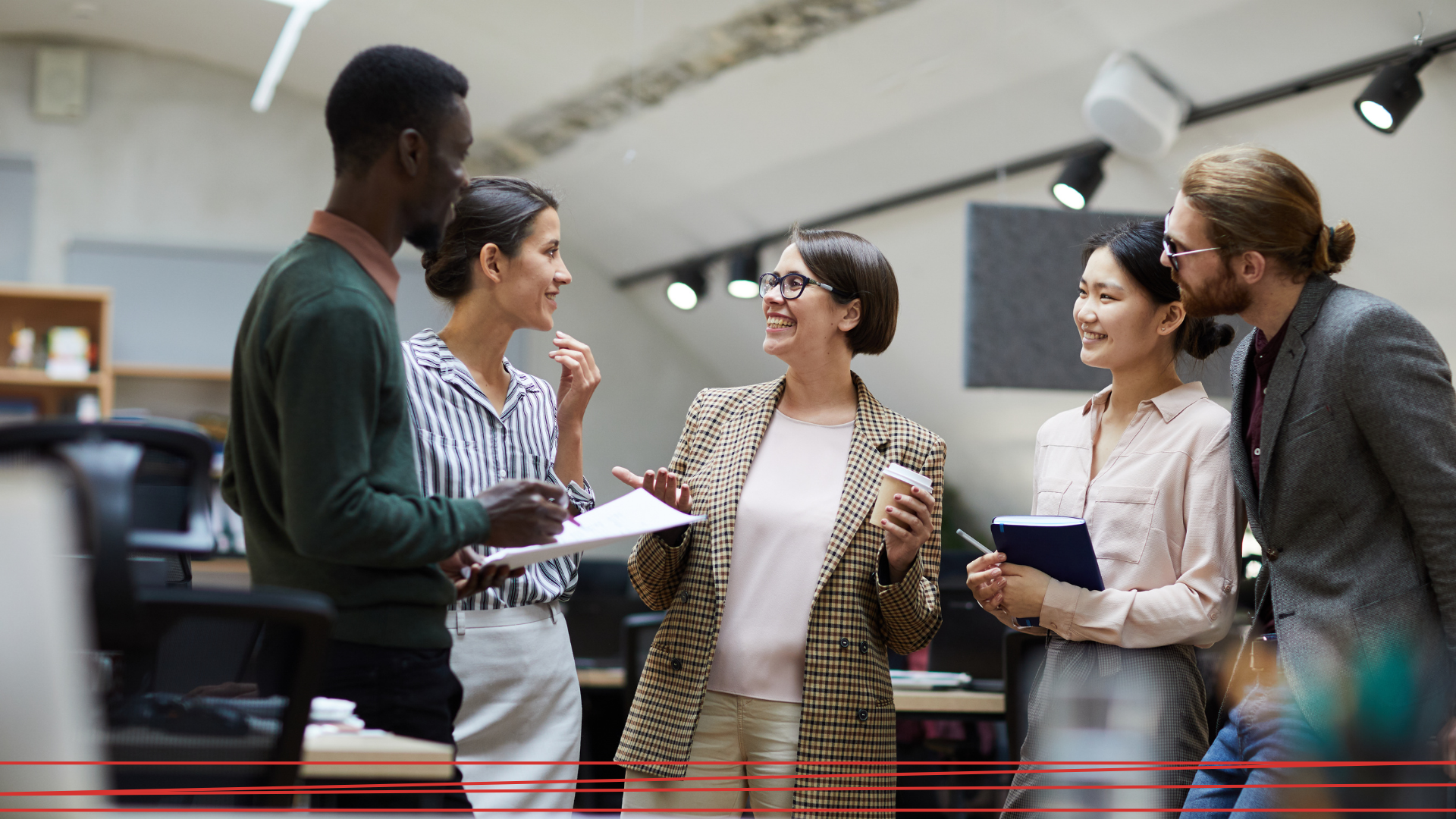 Cinque professionisti in abbigliamento business casual sono in piedi in un ufficio, sorridenti e intenti a conversare, con in mano documenti e tazze di caffè.