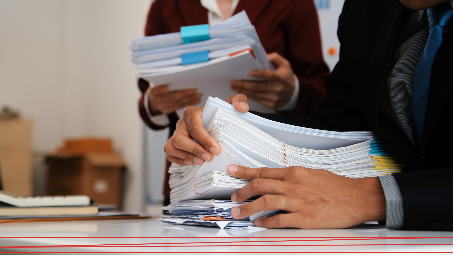 Due persone in abiti da lavoro sistemano grosse pile di documenti cartacei su una scrivania.