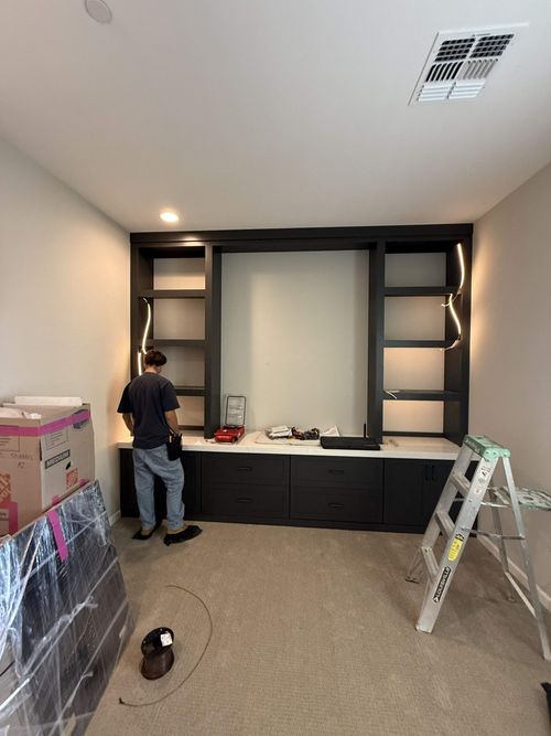 Person assembling dark built-in shelving and cabinets in a room with tools and a ladder.