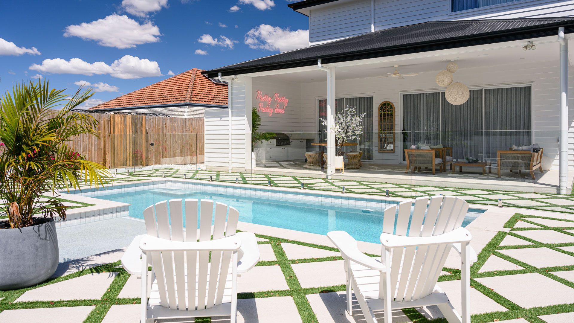 Backyard patio with a pool, white chairs, and a modern house under a blue sky.