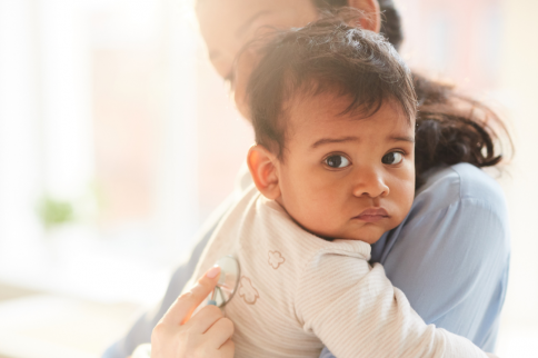 Photo of a small child being held by another person indoors, with the child facing the camera and light colored surroundings visible in the background.