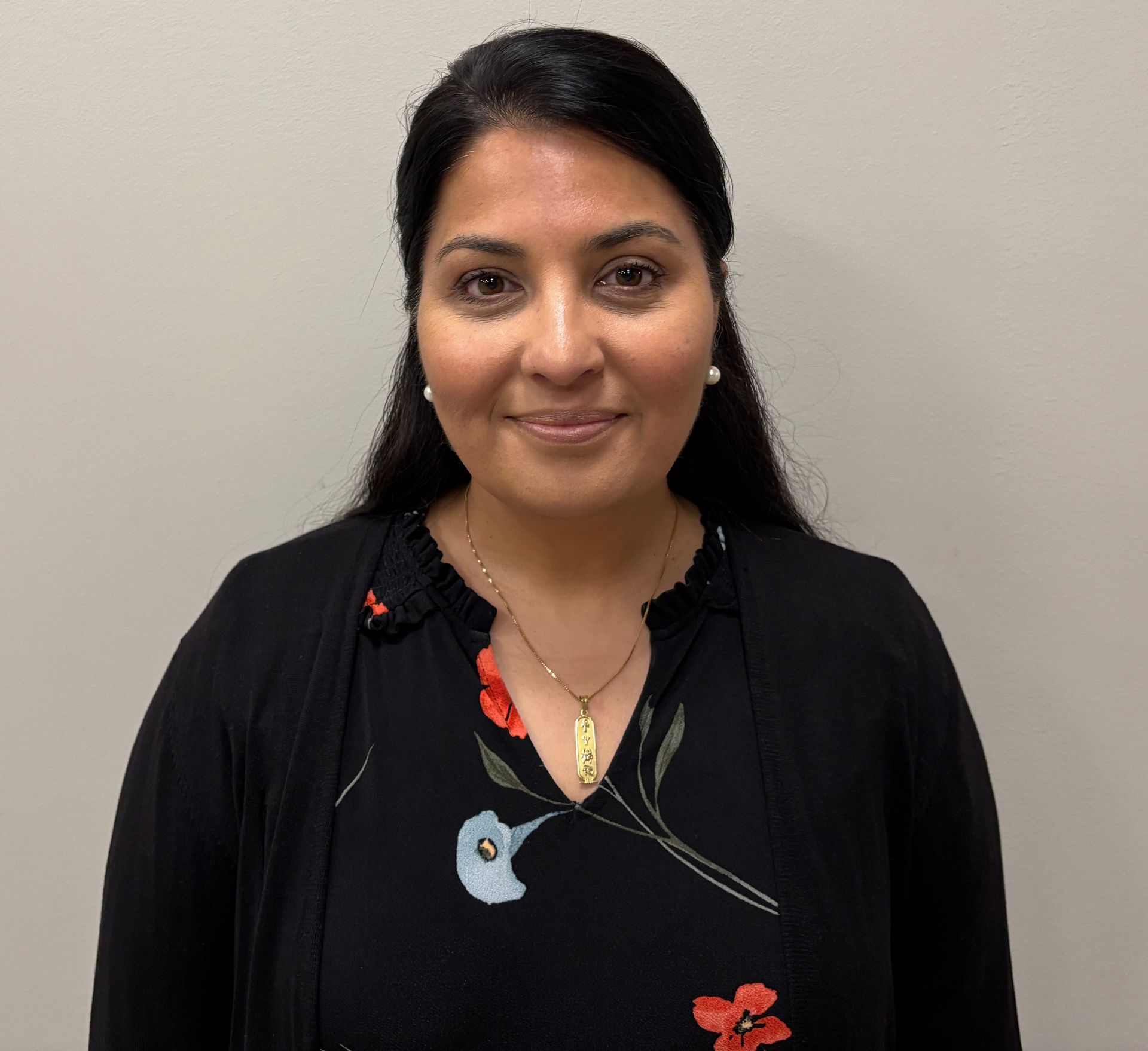 Portrait of a healthcare provider wearing a dark top with a floral design, standing against a plain light colored wall.