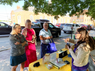 People gathered around a table outdoors, examining small items at a market stall.
