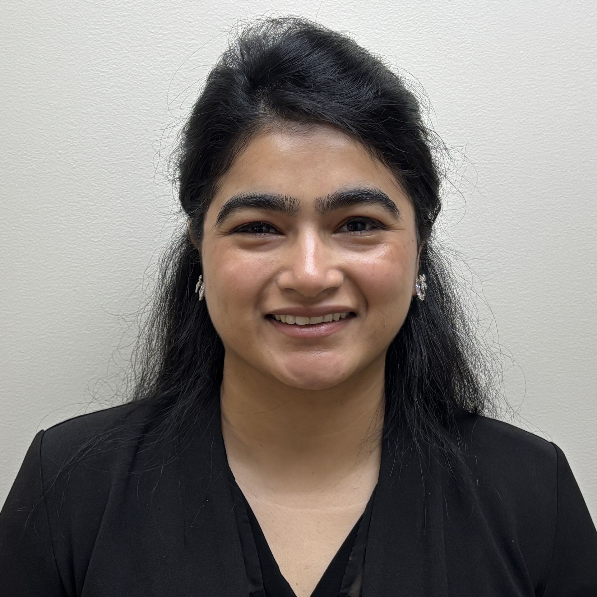 Professional profile photo of Dr. Mehta standing in front of a plain light grey wall wearing a black blouse with her hair up in a half do.