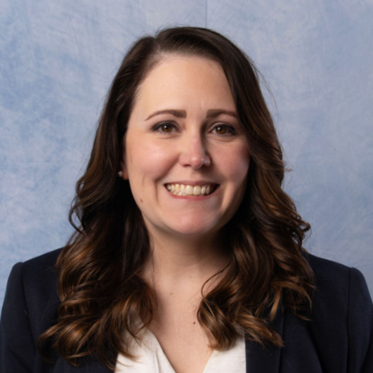 Smiling woman with glasses and brown hair, wearing a blue lanyard against a light wall.
