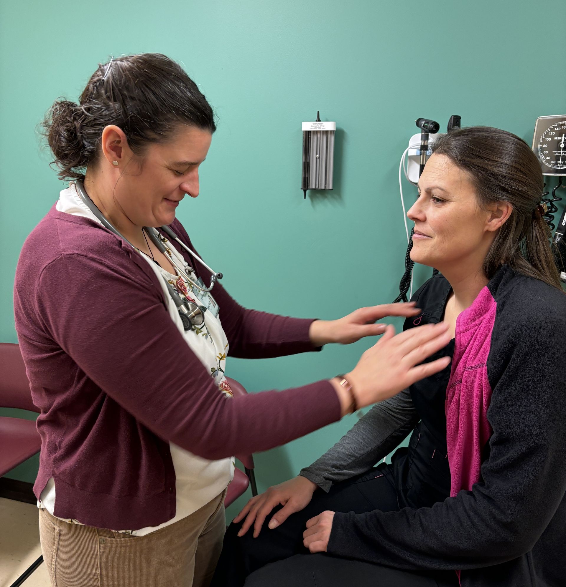 Photo of two people in a medical exam room, with one person standing and placing their hands on the other person’s upper chest to perform an exam while the other person sits on an exam table, with medical instruments and a teal-colored wall visible in the background.