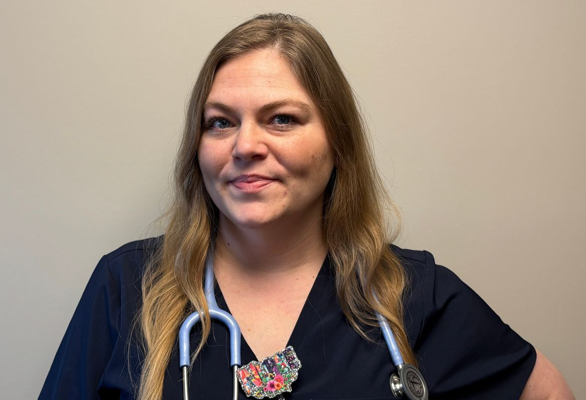 Professional profile photo of Kendra Young, LPN wearing a dark scrub top and a stethoscope, with a decorative badge clipped at the neckline, photographed against a plain light colored background.