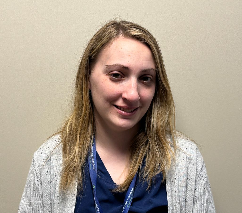 Head-and-shoulders photo of a healthcare professional with shoulder length hair, wearing a dark top, light cardigan, and a lanyard, photographed indoors against a plain light colored wall.