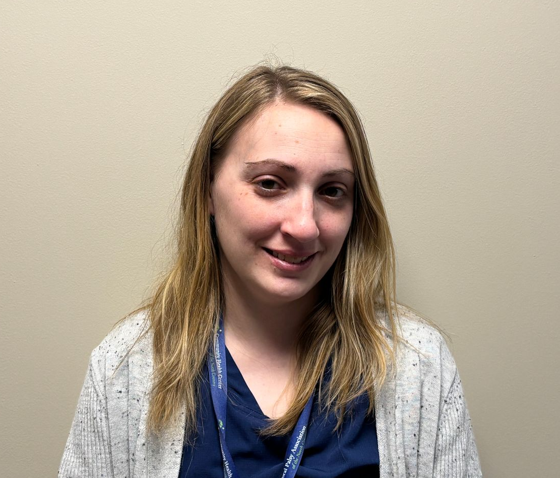 Head-and-shoulders photo of a healthcare professional with shoulder length hair, wearing a dark top, light cardigan, and a lanyard, photographed indoors against a plain light colored wall.