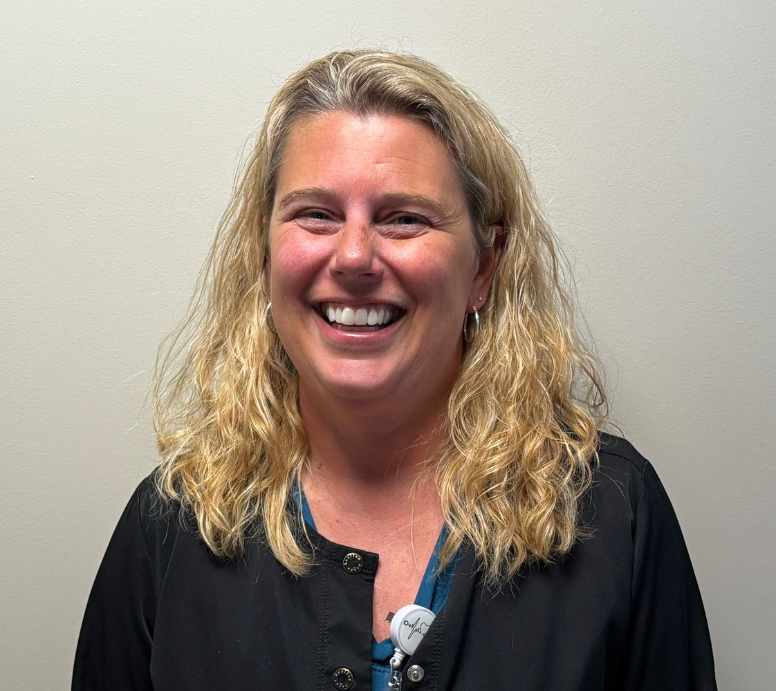 A portrait-style photograph of a person with shoulder‑length wavy blonde hair standing against a plain light-colored wall. They are wearing a dark top layered over a blue shirt, with an identification badge clipped to the front. The badge includes a photo and text, but the details are too small to read