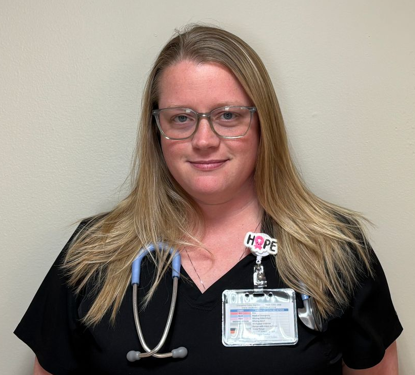Head and shoulders portrait of an adult standing against a plain, light colored wall. The person has long, light colored hair and is wearing clear framed glasses and a black scrub top. A stethoscope is draped around the neck, and an ID badge is clipped to the chest.  The person faces the camera directly.
