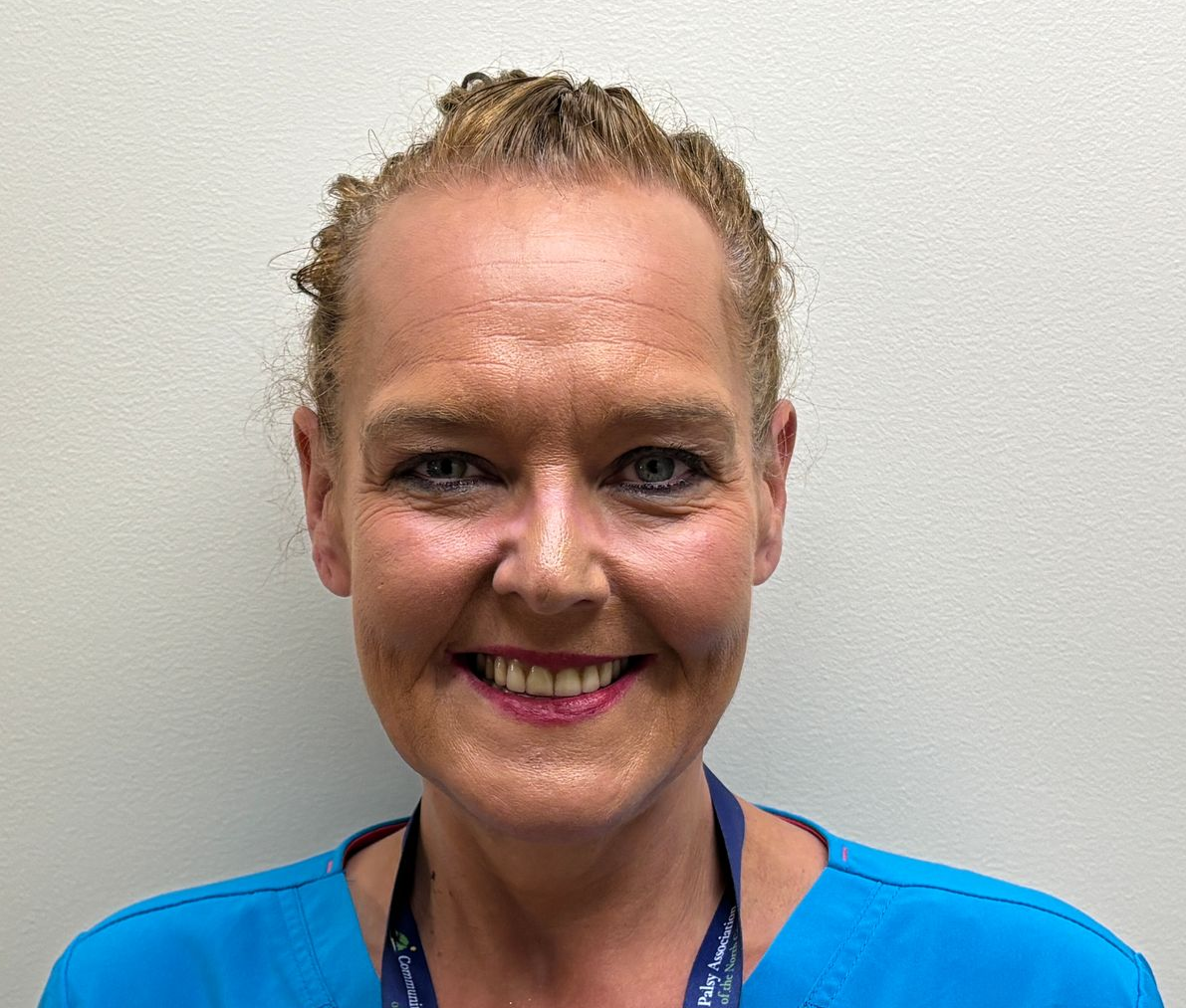Close-up, centered head-and-shoulders portrait of an adult wearing a bright blue scrub top and a dark lanyard with white text and small logos. The person has light-colored hair pulled back and is facing the camera with a visible smile. The background is a plain, light-colored wall with even lighting. The lanyard hangs from the neck and extends out of frame.