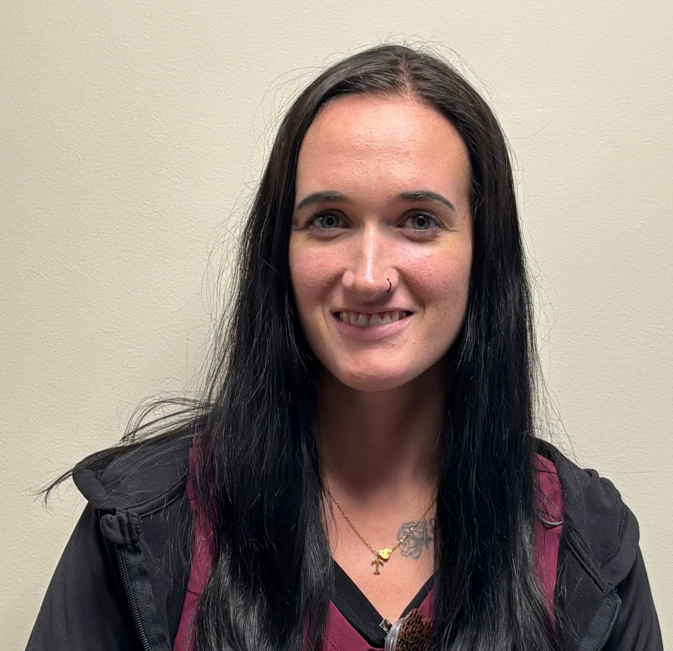 A woman with long dark hair sits against a plain beige wall. She wears a maroon scrub top under a black zip‑up jacket.