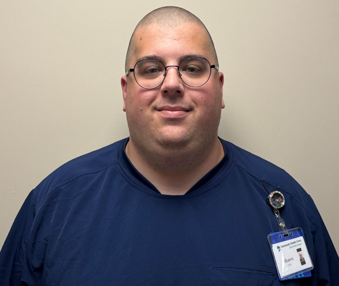 Man in blue shirt with glasses and ID badge, standing against a plain beige wall.