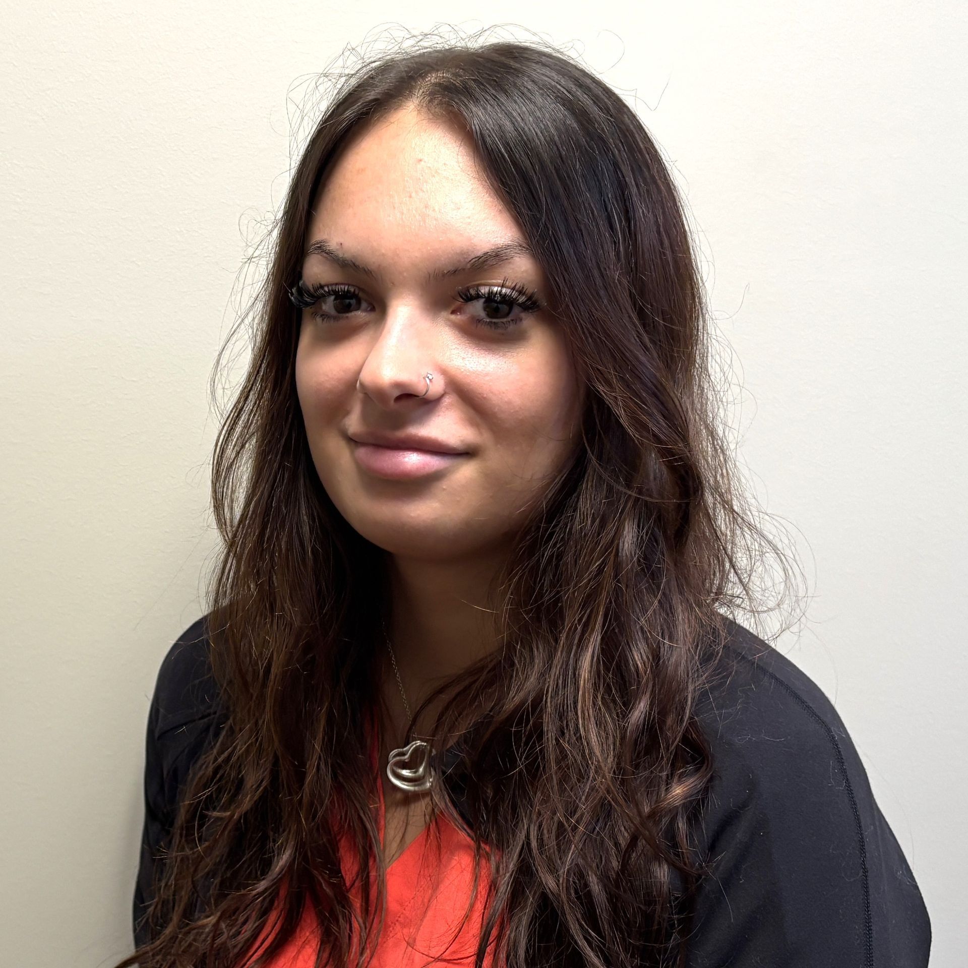 Portrait of a person with long dark hair wearing a pink top, standing against a plain beige wall.