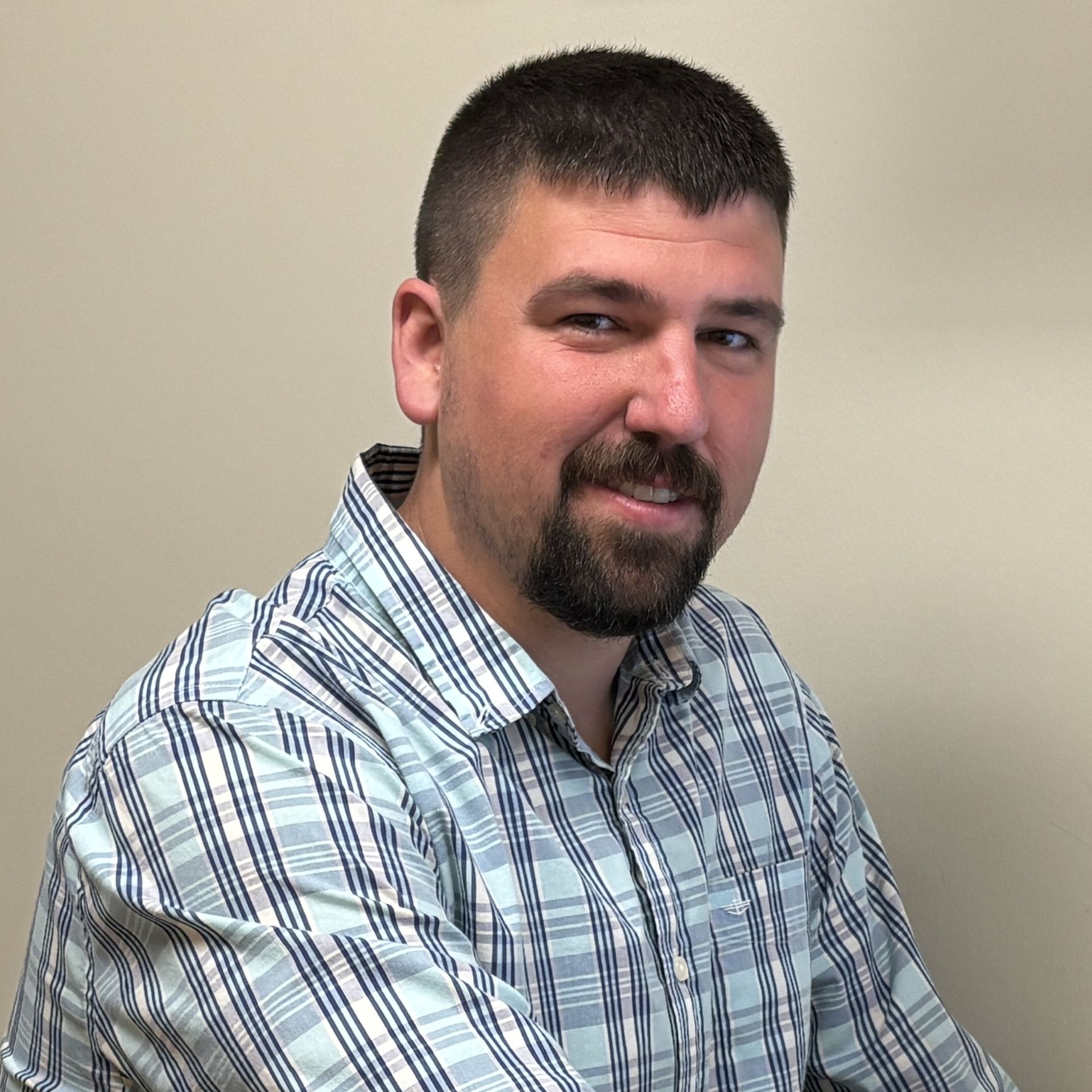 A portrait photograph of a person standing in front of a plain beige wall. The person has short dark hair, a trimmed beard, and is wearing a light blue and white plaid button‑up shirt. The lighting is even, and the image appears to be taken indoors.