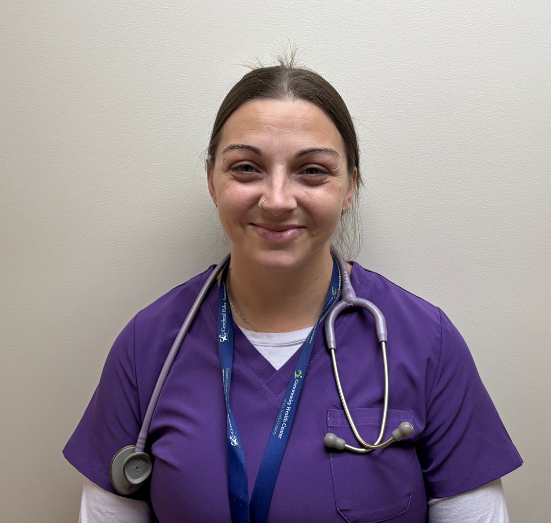 Head-and-shoulders profile photo of Melonie Cooper, Medical Assistant  wearing a purple scrub top, a white long-sleeve undershirt, a lanyard, and a stethoscope, photographed against a plain light-colored background.