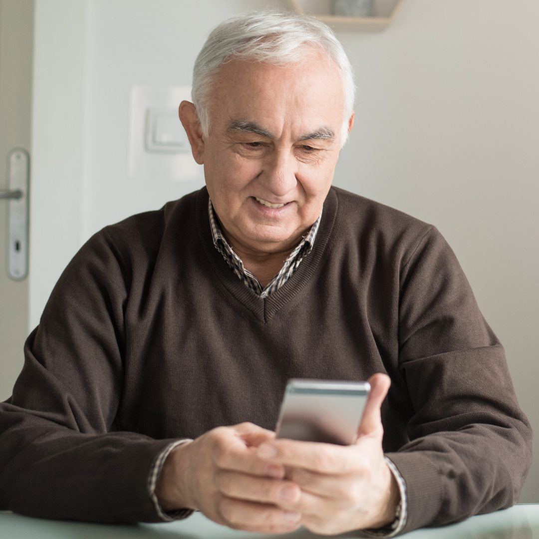 Photo of an older adult seated at a table indoors, wearing a dark sweater over a collared shirt and holding a smartphone, with light colored walls and household fixtures visible in the background.