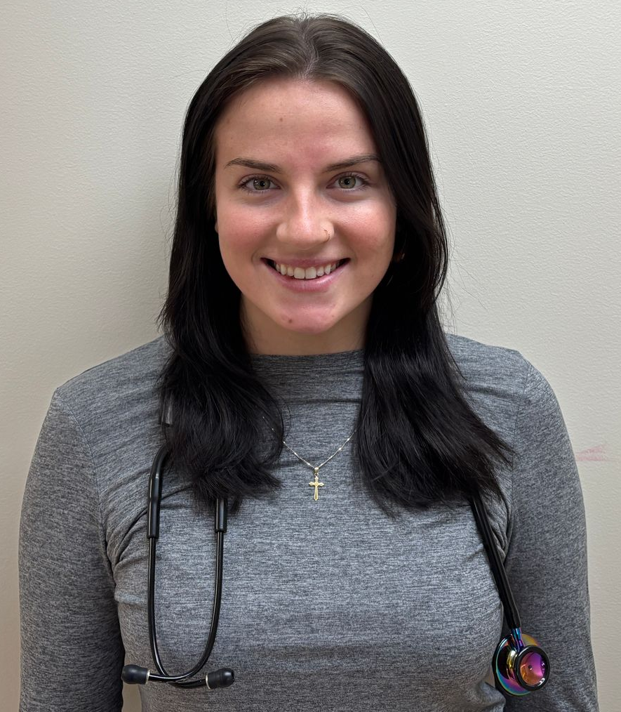 Portrait of Riley Storrin, Medical Assistant wearing a gray long-sleeve top and a stethoscope, standing against a plain light colored background.