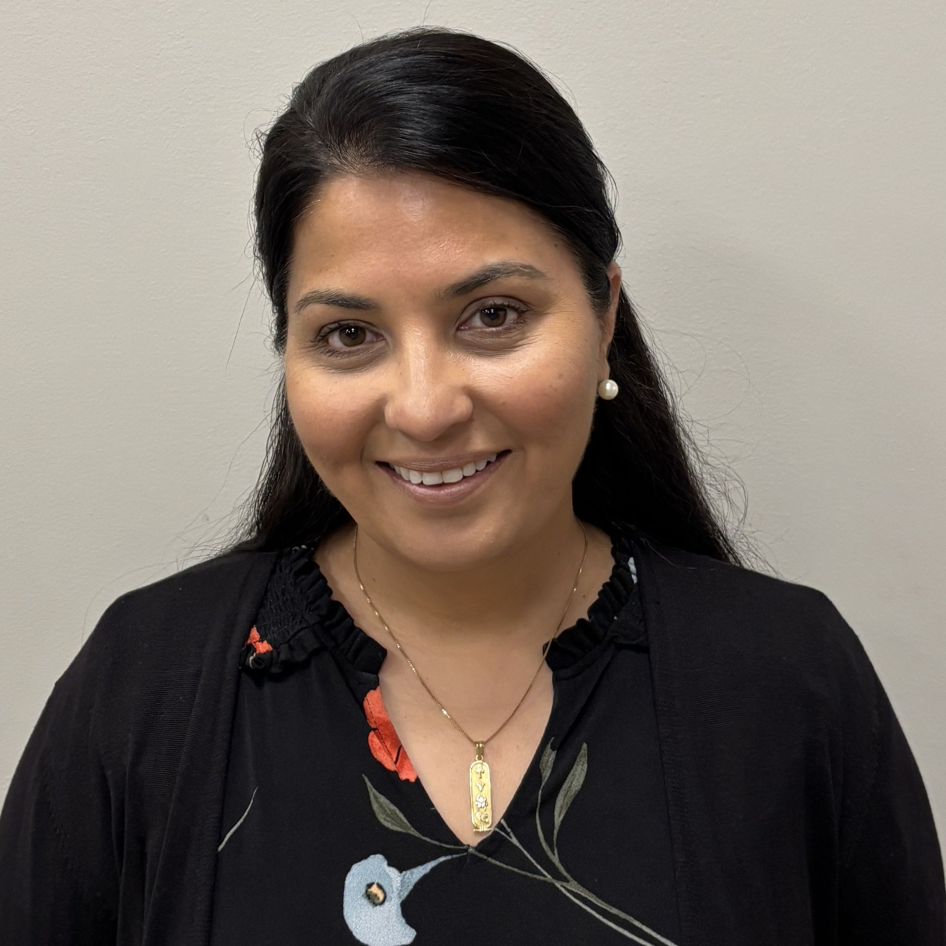 Professional profile photo of Dr, Shah Sekhon, MD wearing a black cardigan over a black floral-patterned top and a necklace, photographed against a plain light colored background.
