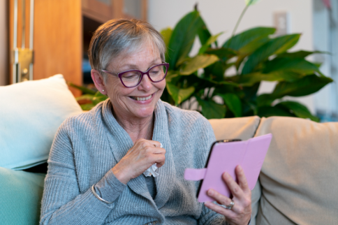 Photo of an older adult seated on a couch indoors, wearing glasses and a gray cardigan, holding a tablet with a pink cover, with houseplants and neutral furnishings visible in the background.