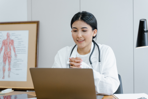 Photo of a person wearing a white medical coat and a stethoscope, seated at a desk and looking at a laptop computer, with a medical poster and office furnishings visible in the background.