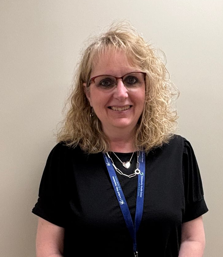 Woman with a badge standing in office hallway, wearing glasses, black shirt, and white patterned skirt.