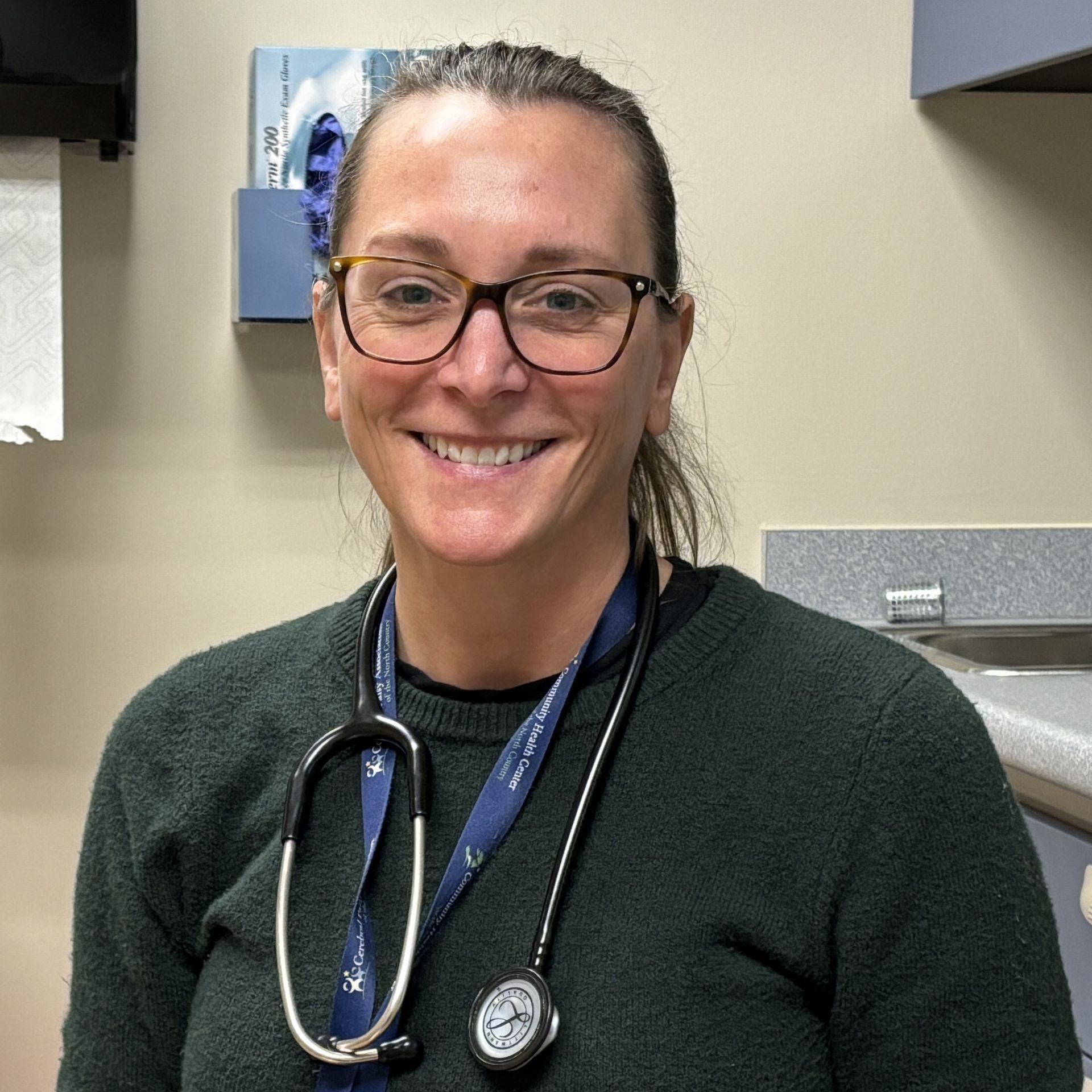 Picture of Adult Nurse Practitioner Christine Thrasher wearing a green sweater with Stethoscope hung around her neck.  Background is an exam room.