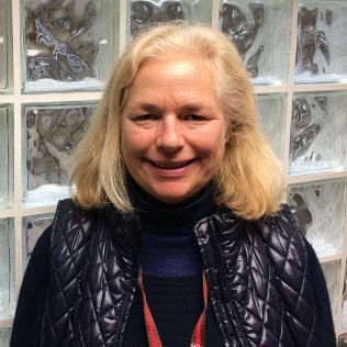 Head-and-shoulders profile photo of Cynthia Wells, Outreach and Enrollment Coordinator wearing a dark quilted vest over a dark top and a lanyard, photographed indoors in front of a glass block wall.