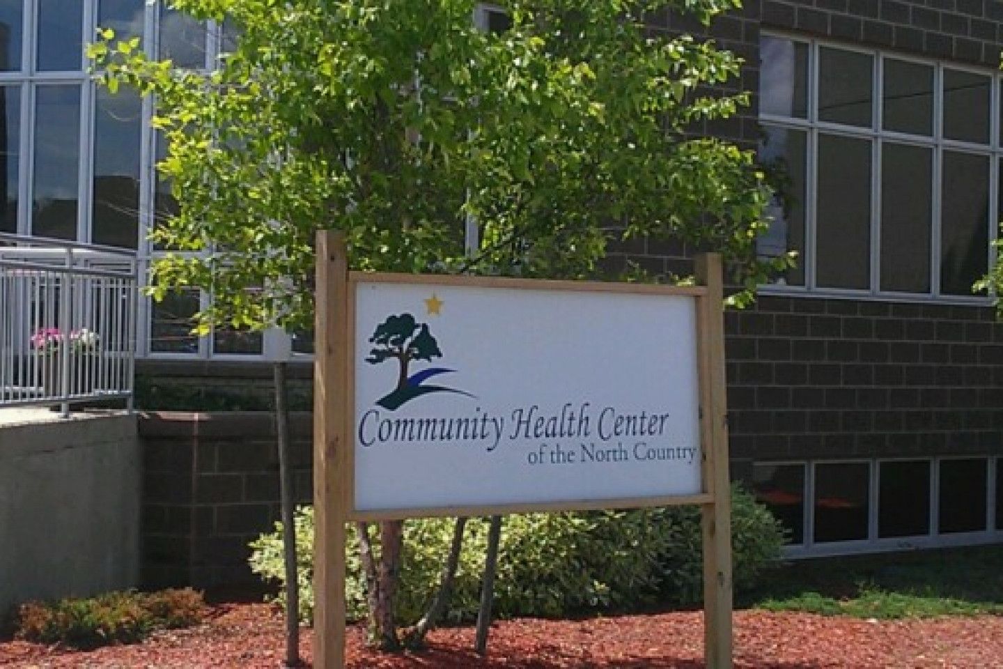 Exterior photo of a freestanding sign at the entrance to our health center in Gouverneur reading “Community Health Center of the North Country,” mounted on wooden posts in front of a building with trees, landscaping, and windows visible in the background.
