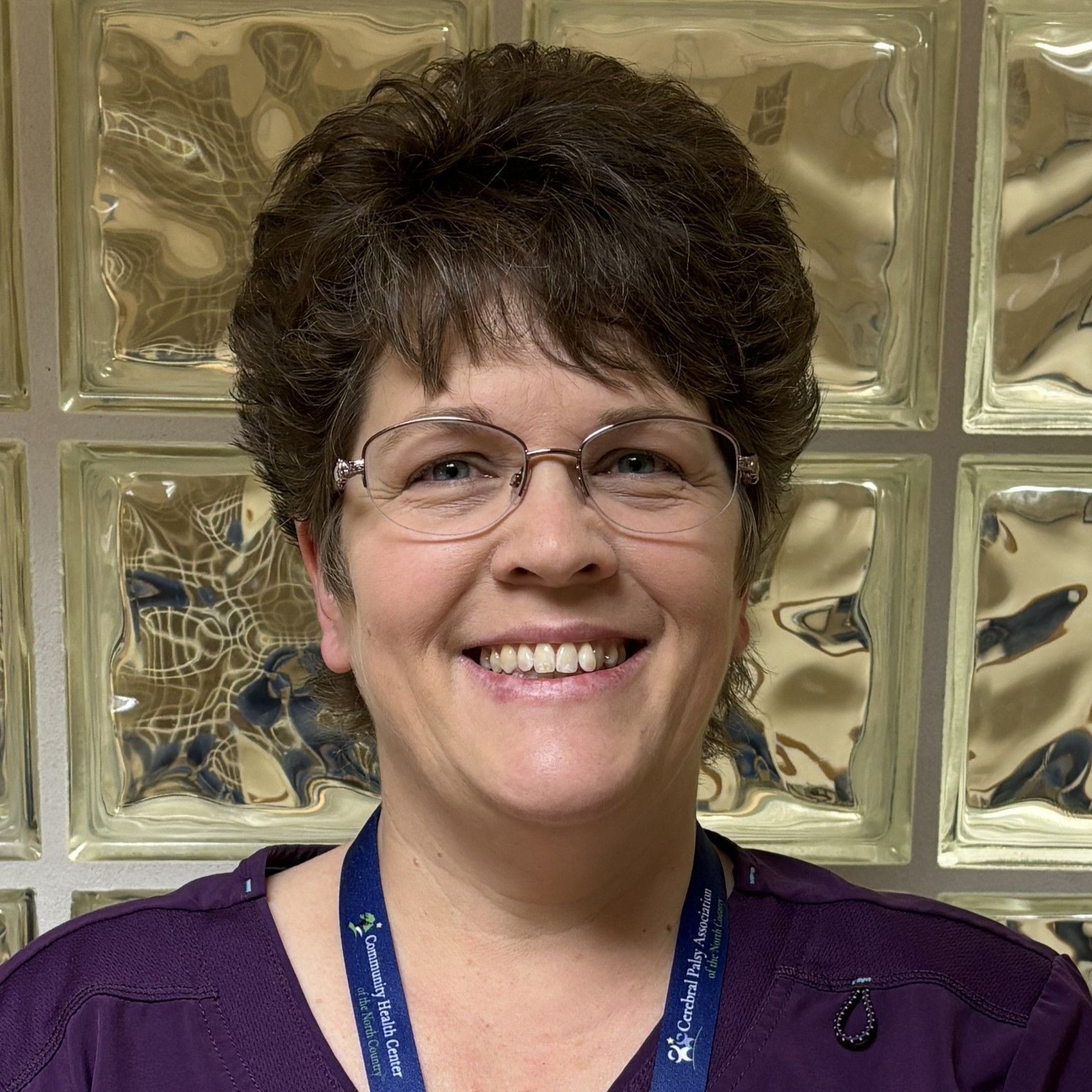 Professional profile photo of Ami Cole, LPN wearing glasses, a purple top, and a lanyard, photographed indoors in front of a glass block wall.