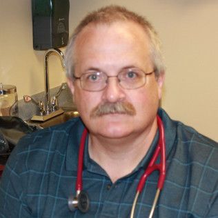 Head-and-shoulders profile photo of Ernie Couglar, Family Nurse Practitioner  wearing glasses, a teal patterned collared shirt, and a red stethoscope, photographed indoors with medical equipment visible in the background.