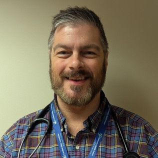 Professional photo of Dr. Scott Spicer, MD wearing a plaid button-down shirt, a lanyard, and a stethoscope, photographed against a plain light-colored background.  Member of the pediatric team at the Gouverneur Health Center.
