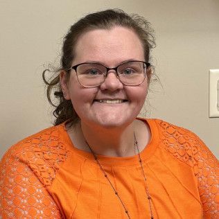Profile photo of Nicole Bonno, Family Nurse Practitioner wearing glasses, an orange lace-textured top, and a necklace, photographed against a plain light-colored background.