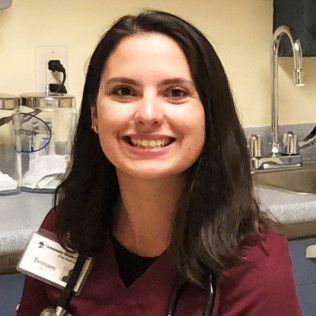 Professional profile photo of Brittany Terpstra, Physician Assistant wearing a maroon scrub top, a name badge, and a stethoscope, photographed indoors with medical supplies and an exam room sink visible in the background.