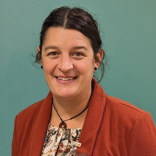 Professional profile photo of Abigail Smith, Family Nurse Practitioner wearing an orange jacket over a patterned top and a necklace, photographed against a plain teal-colored background.