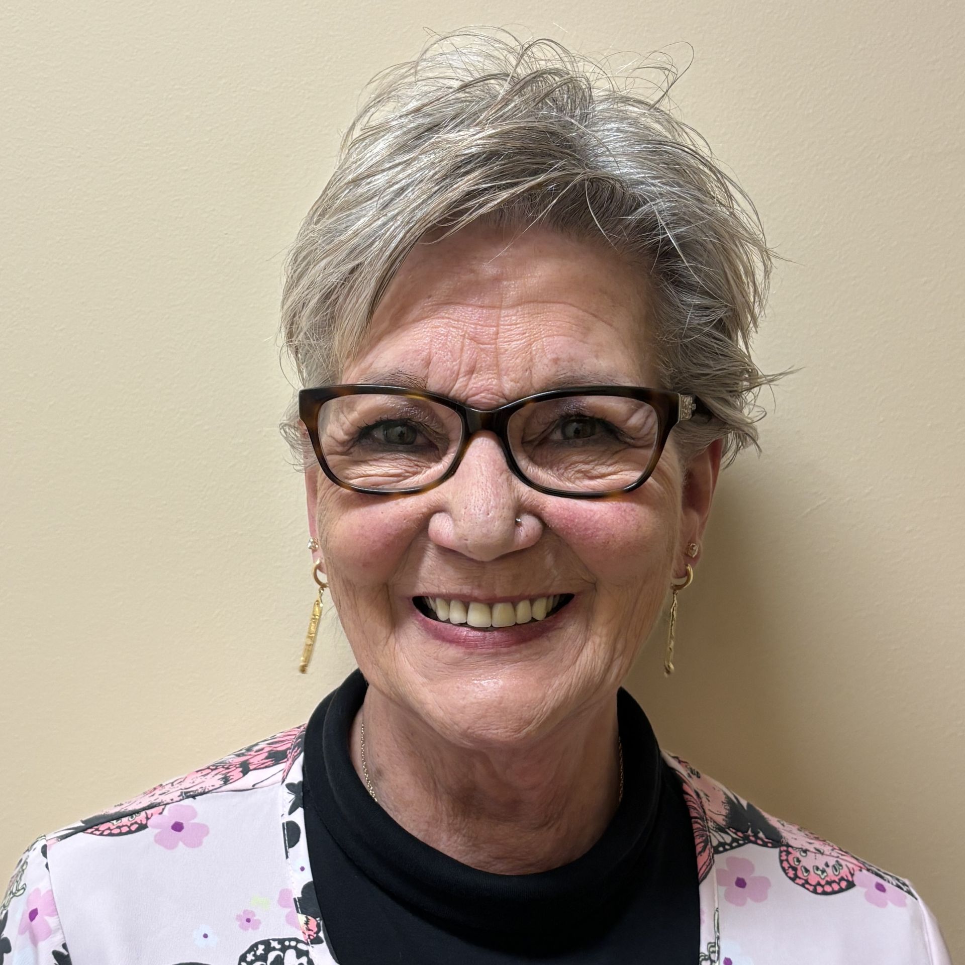 Head-and-shoulders profile photo of Mary Barcomb, Medical Assistant wearing glasses, a black top, and a light-colored floral-patterned jacket, photographed against a plain light-colored background.