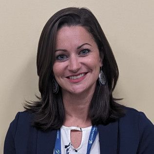 Professional profile photo of Alyson Felt, Certified Family Nurse Practitioner wearing a navy blazer over a light-colored patterned top, along with earrings and a lanyard, photographed against a plain beige background.