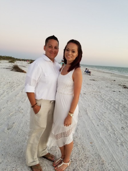 Two people standing together on a white sandy beach at sunset, smiling toward the camera.