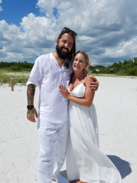 Couple in white outfits smiling on a bright sandy beach under a blue sky