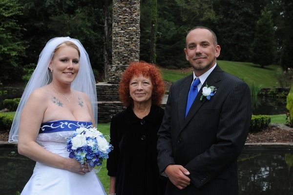 Wedding couple posing with an officiant outdoors beside a pond, bride holding a blue bouquet.