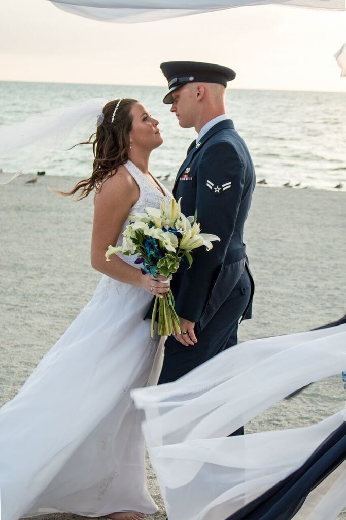 Bride and groom embrace by the sea, holding a bouquet in flowing wedding attire.
