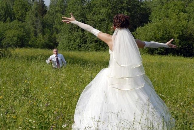 Bride in a white gown with arms outstretched in a grassy field, facing a man in the distance.