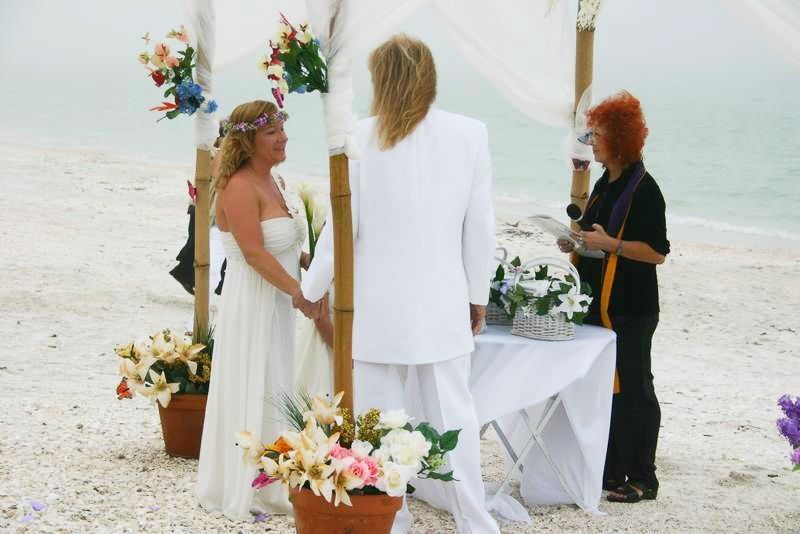 Beachside wedding ceremony with officiant and two people beside a flower-decorated arch