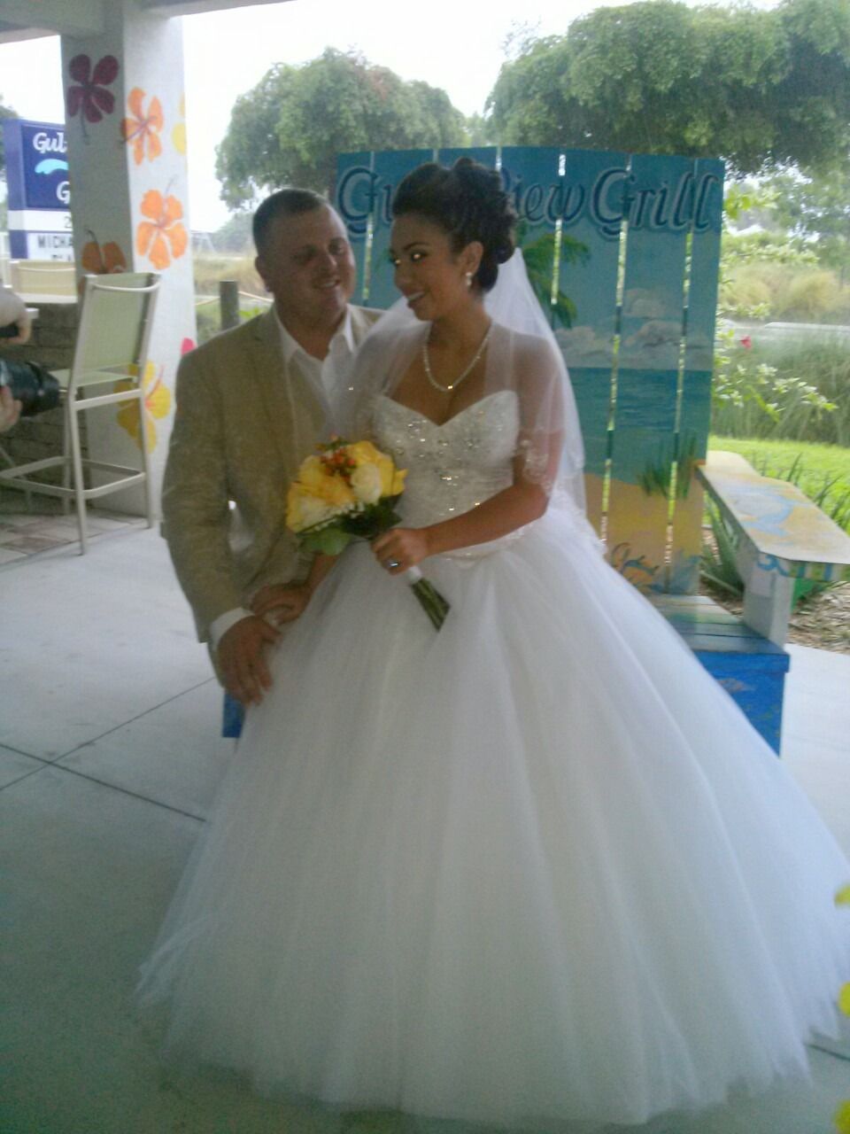 Bride in white gown holding yellow bouquet beside a man at an outdoor wedding ceremony