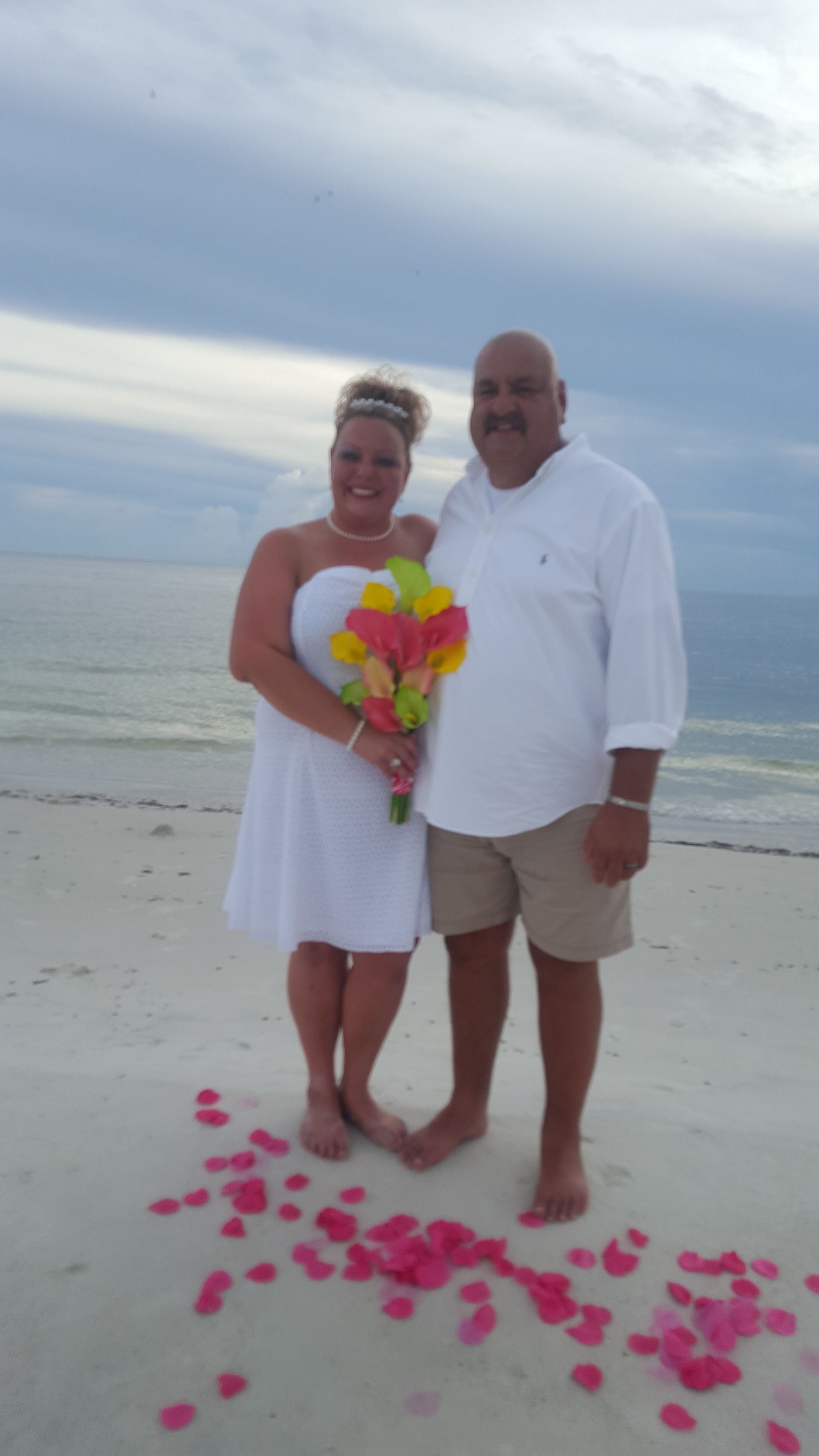Couple standing on a beach in white outfits, holding a colorful bouquet with pink petals scattered at their feet.