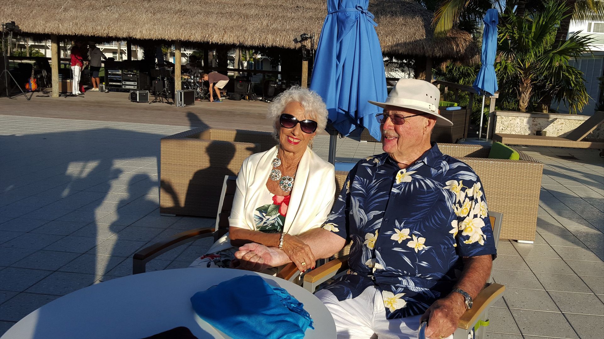 Two people seated at an outdoor café table in sunlight, wearing sunglasses and hats, with a blue umbrella behind them.