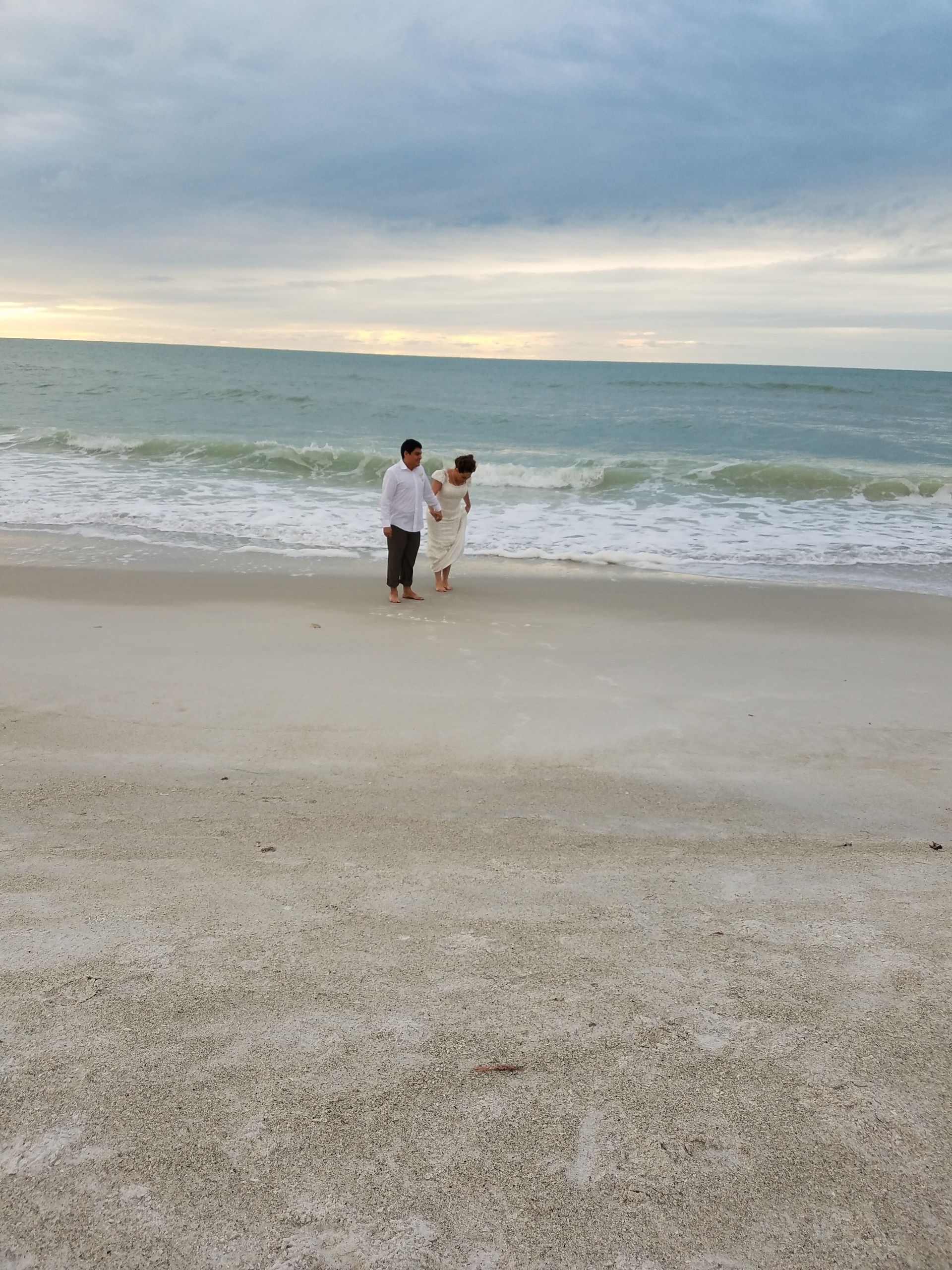 Two people standing on a sandy beach facing the ocean under a cloudy sky