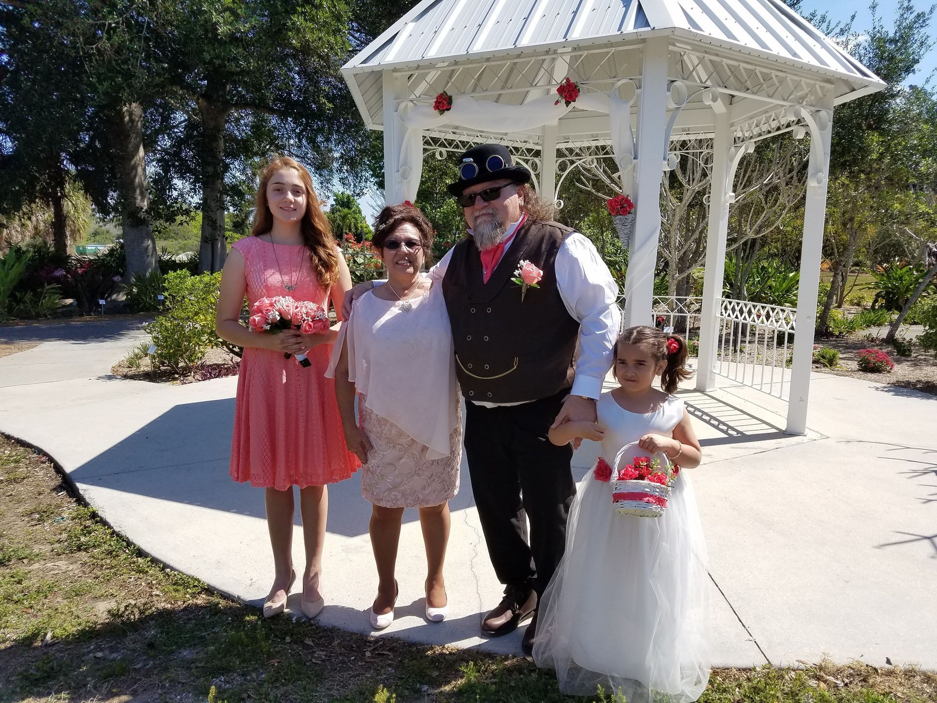 Four people posing in front of a white gazebo outdoors, one in a pink dress and one in a white dress